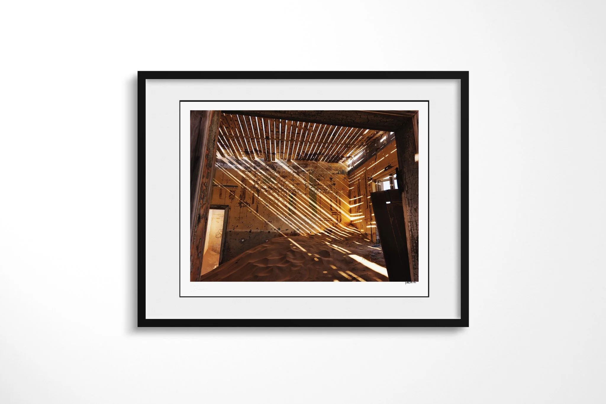 The Ministers House presented in a black frame with a white mount, depicting a sand-filled ochre room crossed by diagonal sunlight from a damaged slatted roof, with peeling walls and an open doorway.