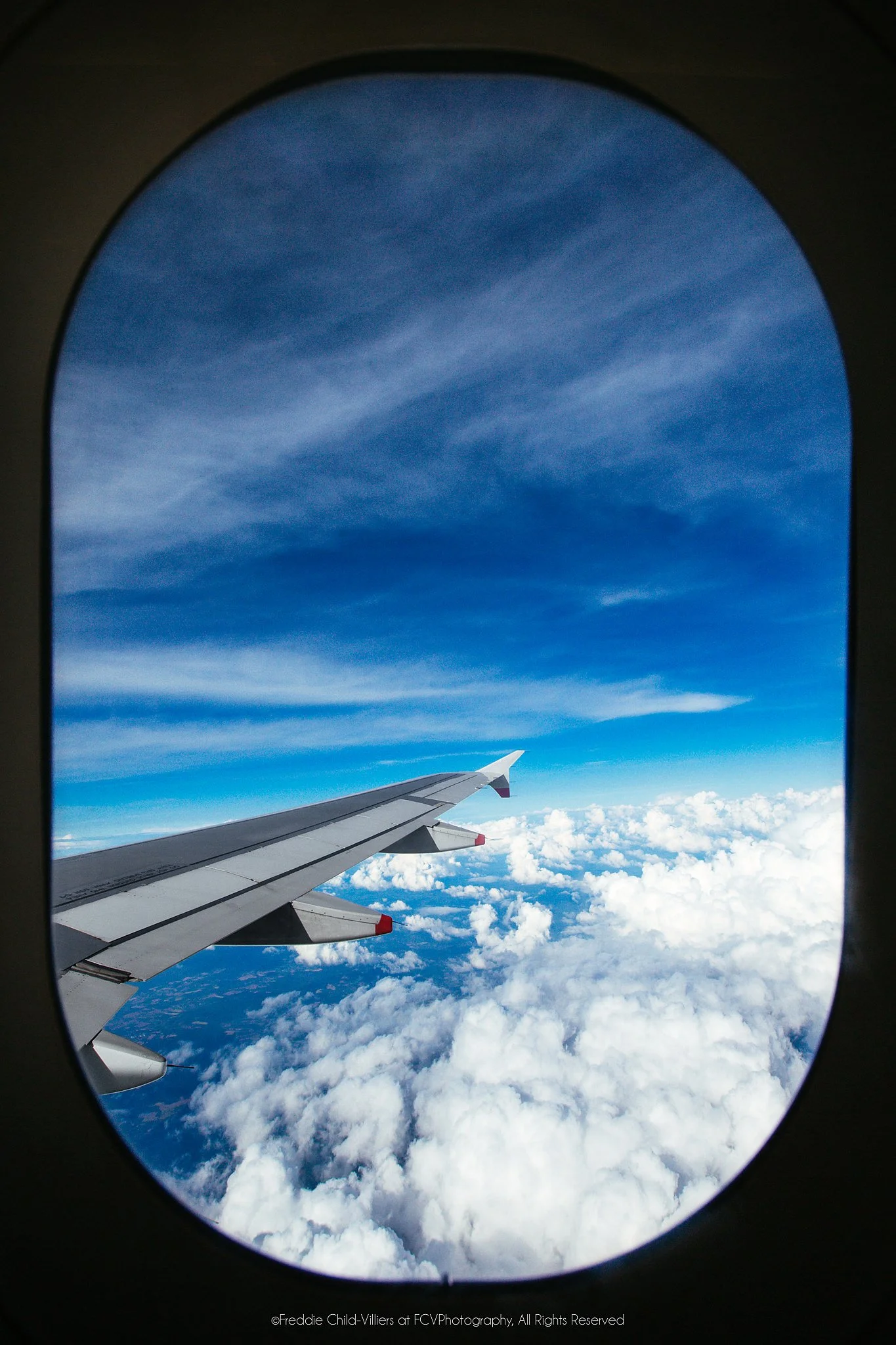 Aircraft wing above bright cloud tops and deep blue sky, framed by the dark oval of a passenger window.