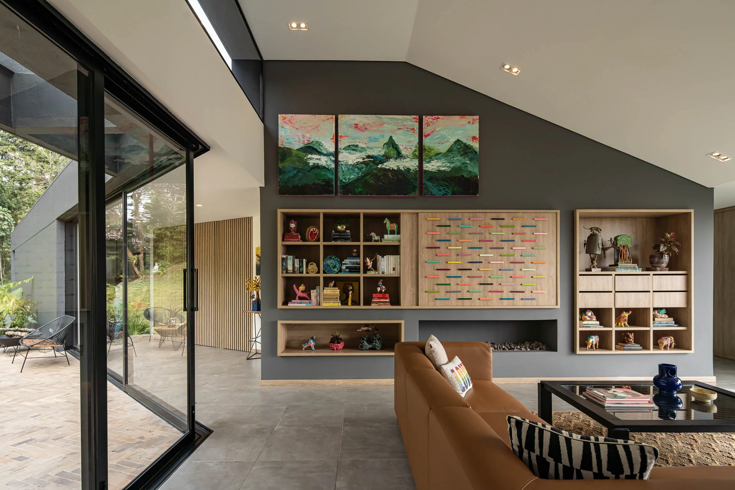 Sala de estar moderna con estanterías de madera, una pintura abstracta de montañas en la pared, sofá marrón y mesa con objetos decorativos, y vista a un patio exterior con sillas y plantas. Casa JANA.