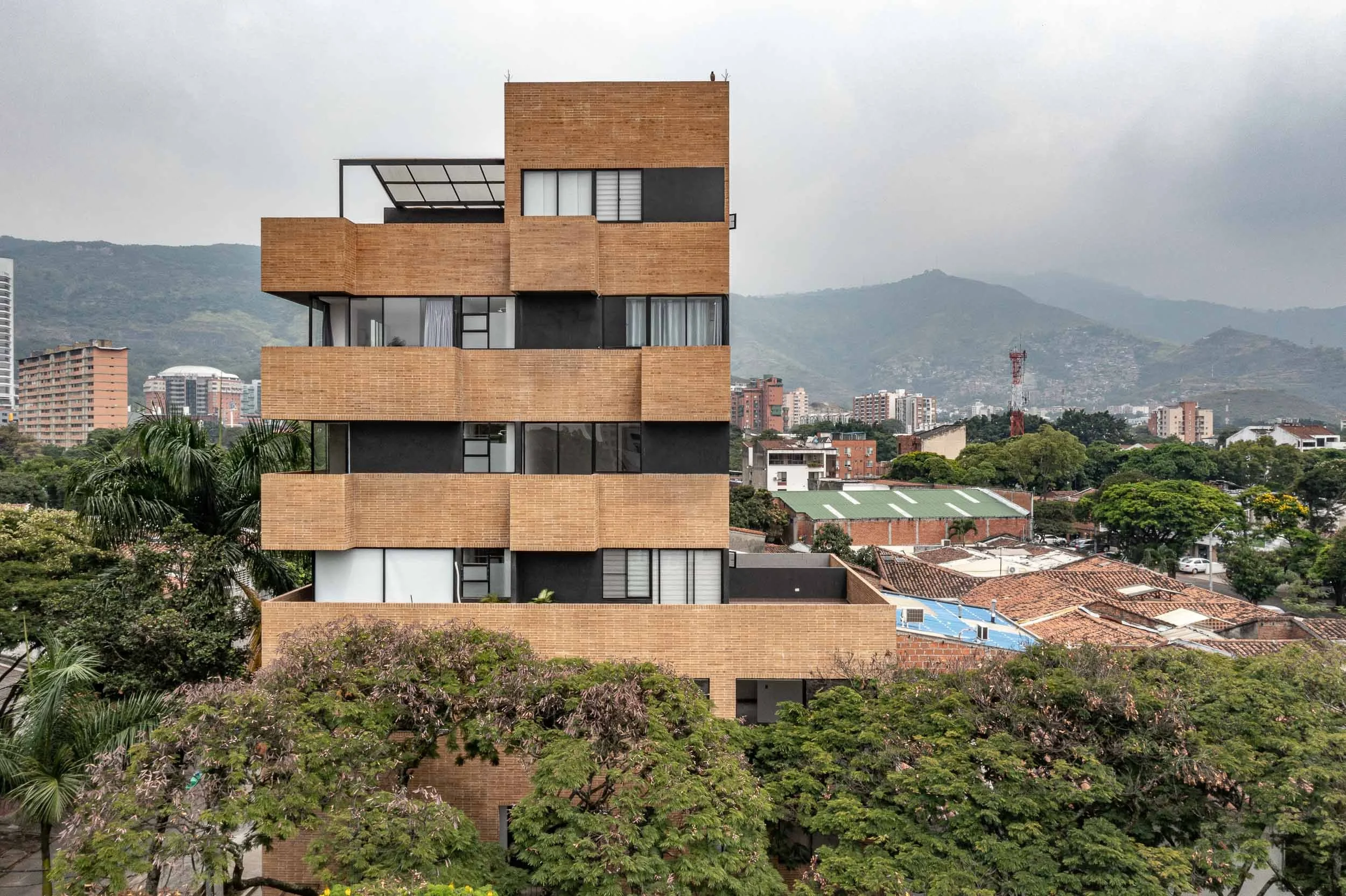 Edificio moderno de varias plantas con balcones de ladrillo y ventanas grandes, rodeado de vegetación en un área urbana con colinas y otros edificios en el fondo. Liv37, edifico moderno en Cali, diseñado por TOLEDO Estudio de Arquitectura.