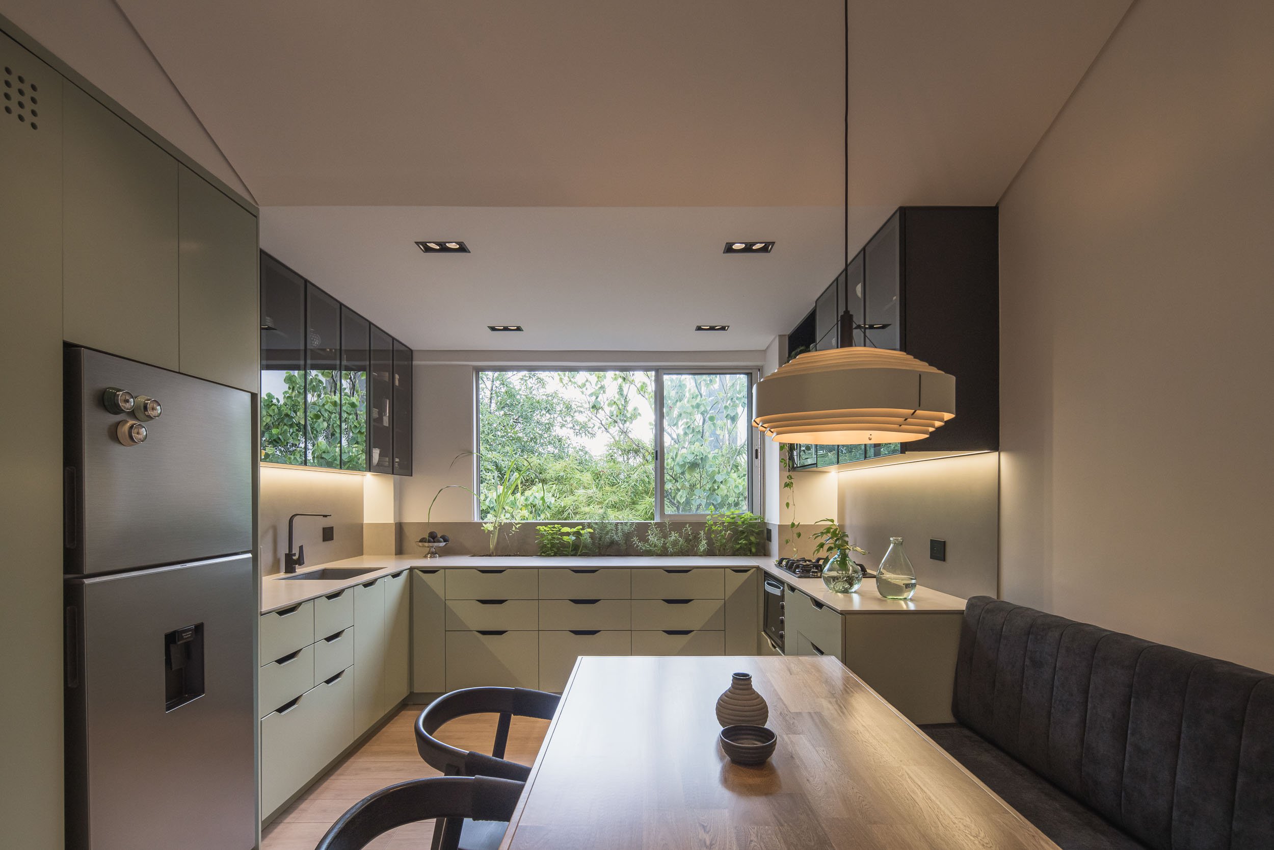 Cocina moderna con gabinetes en tonos verdes y negros, gran ventana con vista a vegetación, mesa de madera y lámpara colgante de diseño. Reforma por TOLEDO Estudio de Arquitectura en El Poblado en Medellín, Colombia.