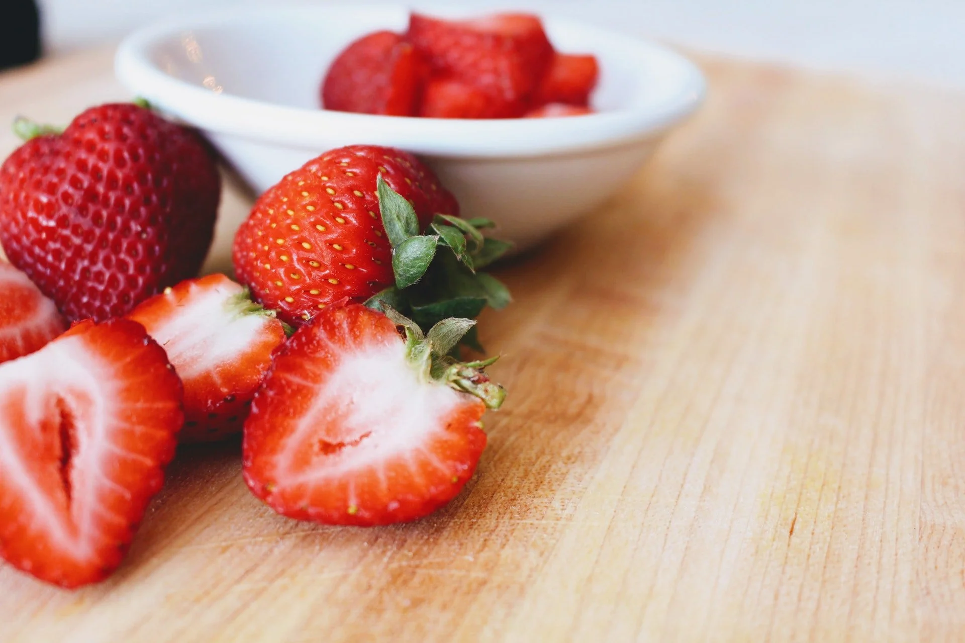 Cut and whole strawberries spilling out of a white bowl onto wood counter