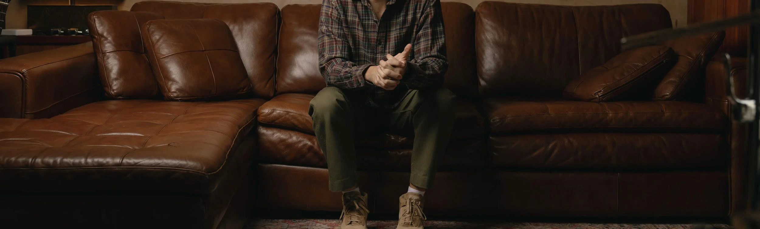 Person sitting on a brown leather couch, wearing a plaid shirt, olive green pants, and tan shoes, with their hands clasped together.
