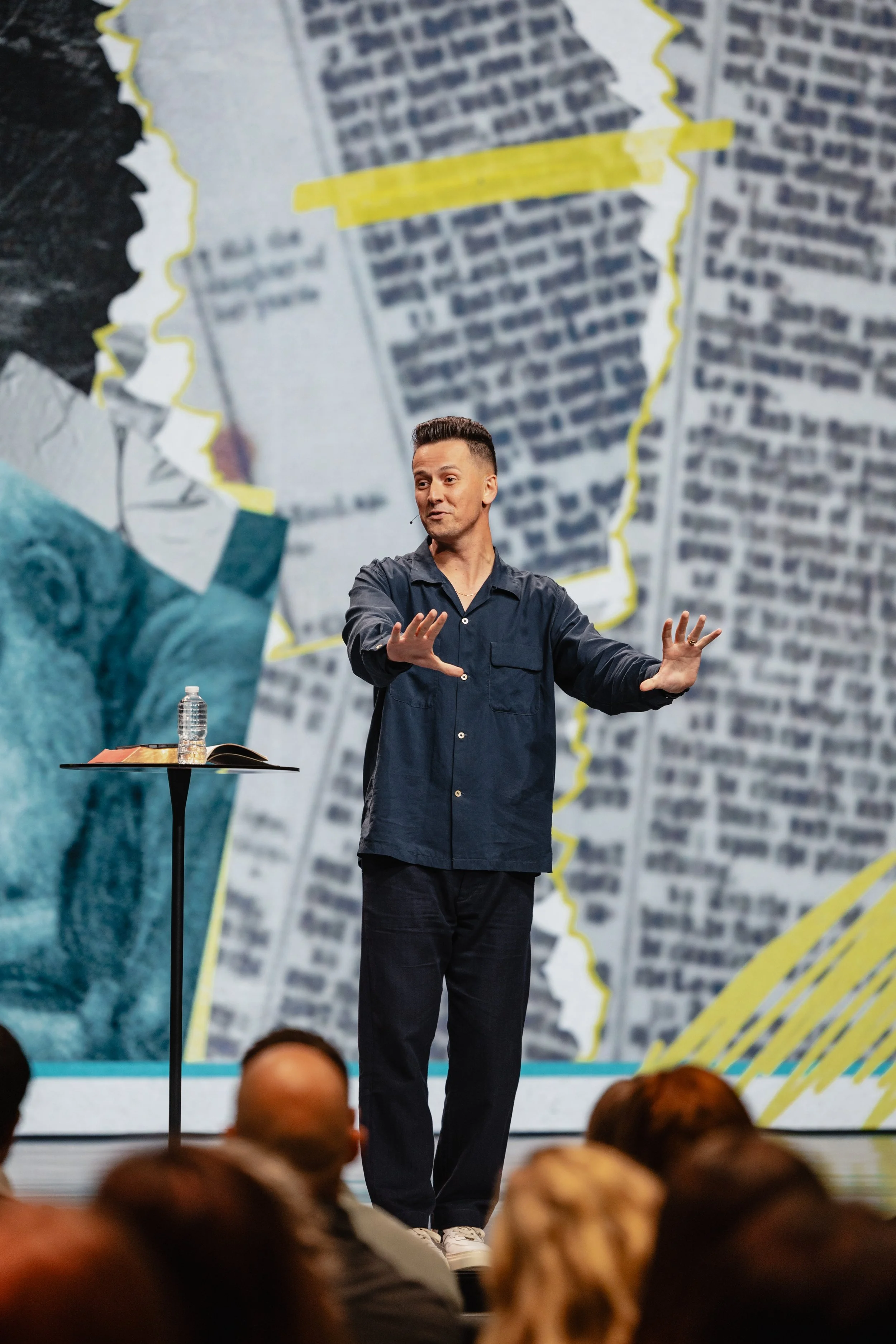 A man standing on stage giving a presentation in front of a large, projected background with text and illustrations. He wears a dark button-up shirt and dark pants, and is gesturing with his hands. A small table next to him holds a bottle of water and an open book. Audience members are visible in the foreground.