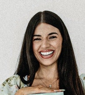 A woman with long dark hair smiling and holding a book, wearing a floral top and a cross necklace.