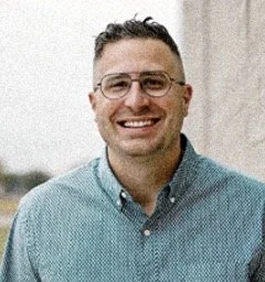A smiling man wearing glasses and a teal button-up shirt standing outdoors with a blurred background.
