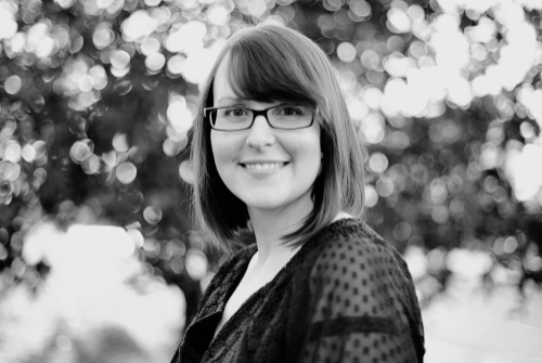 A woman with glasses and shoulder-length hair smiling outdoors with trees in the background.
