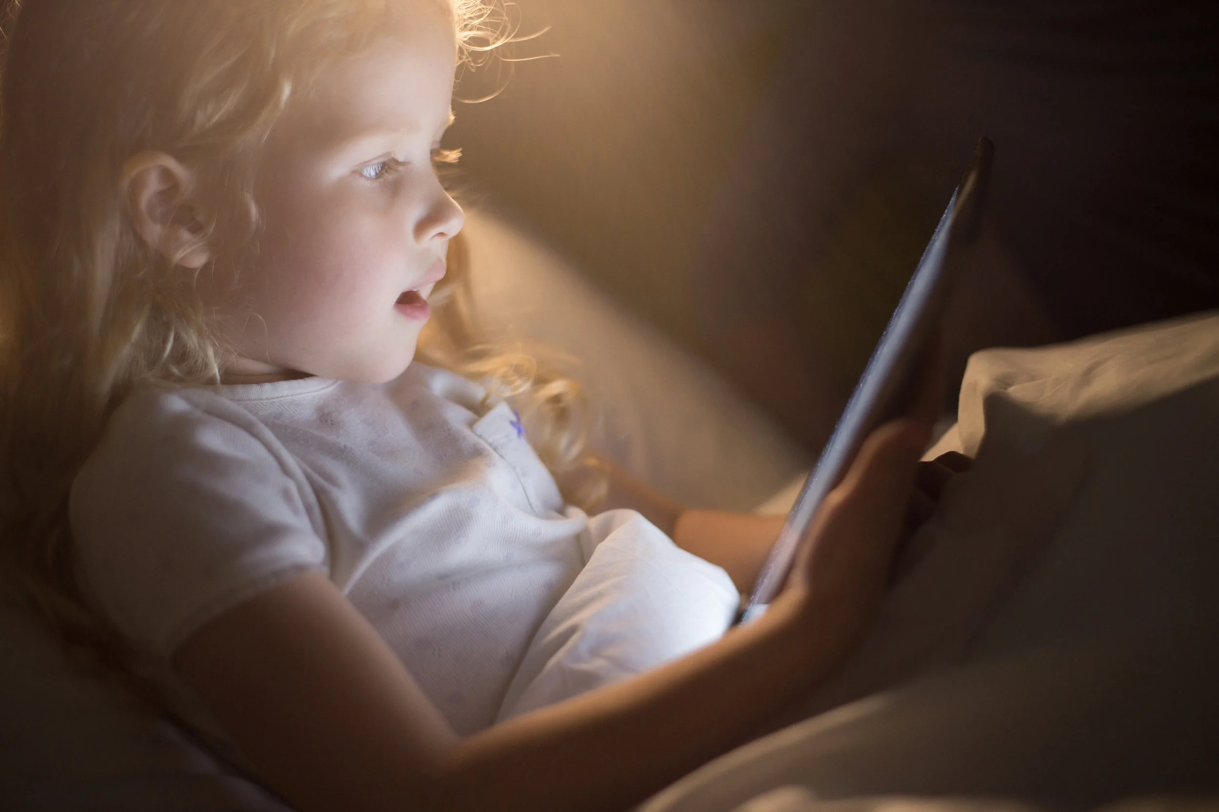 Side_view_portrait_of_adorable_little_girl_looking_amazed_with_mouth_open_using_digital_tablet_lying_in_bed_at_night,_face_lit_by_screen_light_like_magic,_copy_space.jpeg