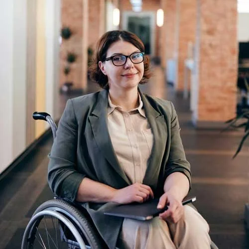 Professional woman seated in a wheelchair in a modern office hallway, wearing glasses and a blazer, holding a tablet and looking at the camera.