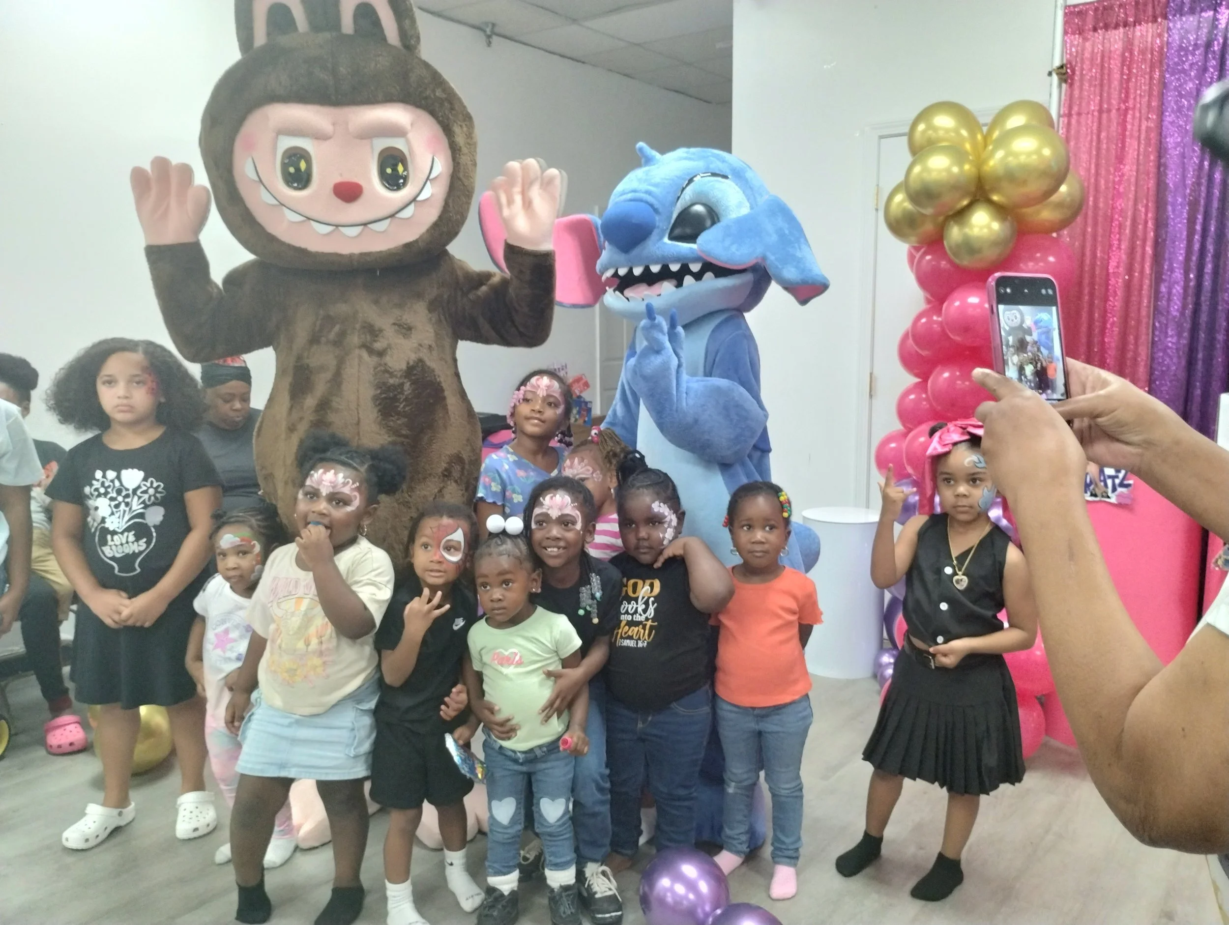 Children at a birthday party with two costumed characters, one in a lion costume and the other in a blue creature costume, taking a group photo.