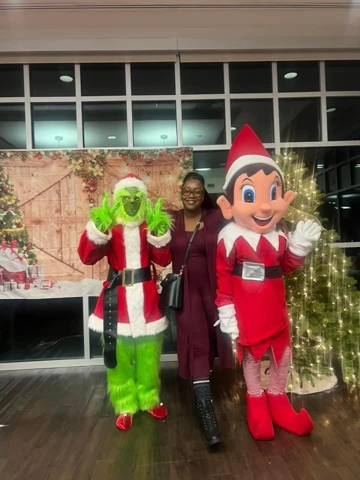 A woman standing between a person dressed as the Grinch and a large Pinocchio character at a Christmas-themed event with a decorated tree and festive backdrop.