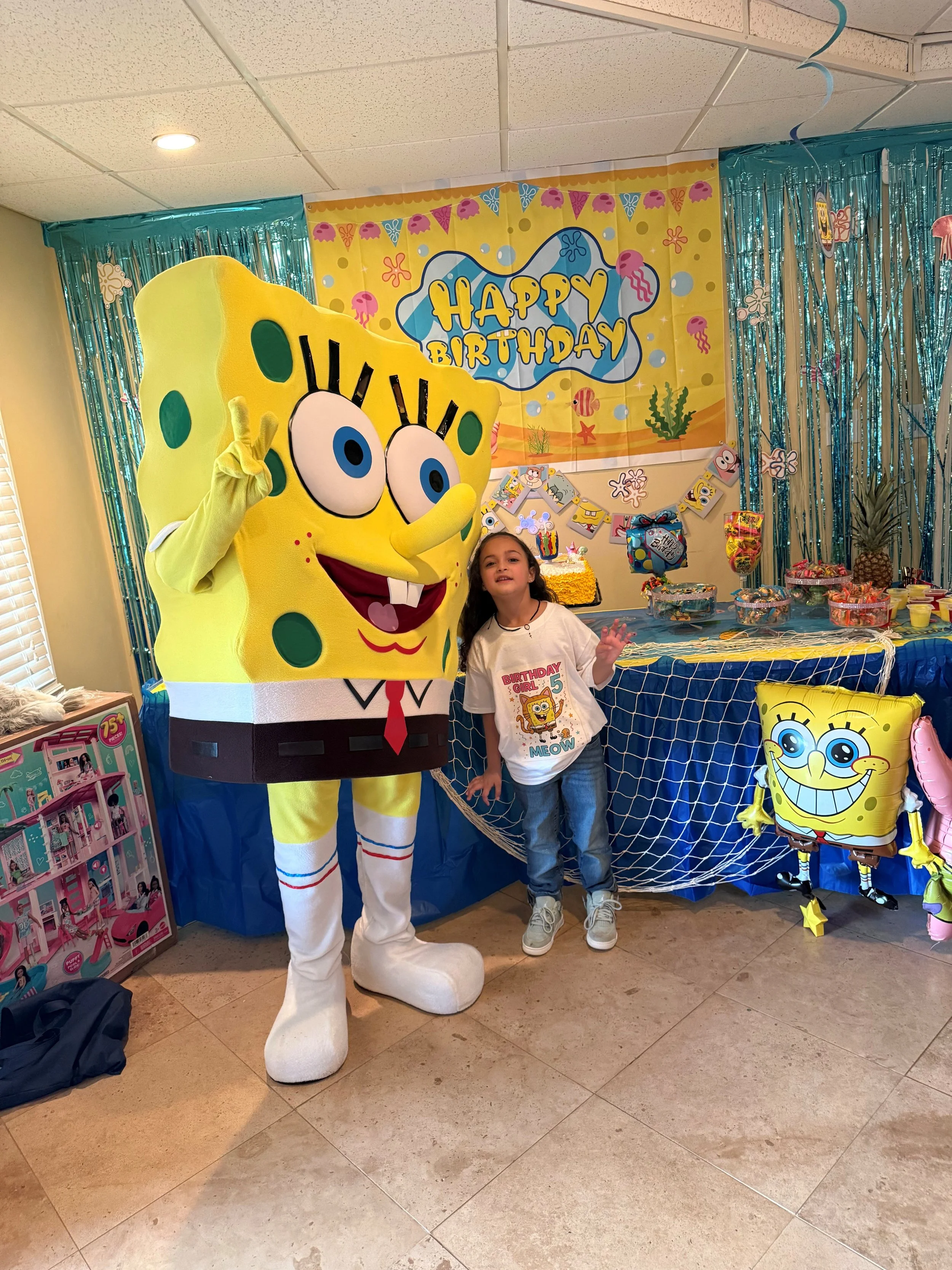 A girl standing next to a person dressed as SpongeBob SquarePants at a birthday party with a decorated table, balloons, and a 'Happy Birthday' banner in the background.