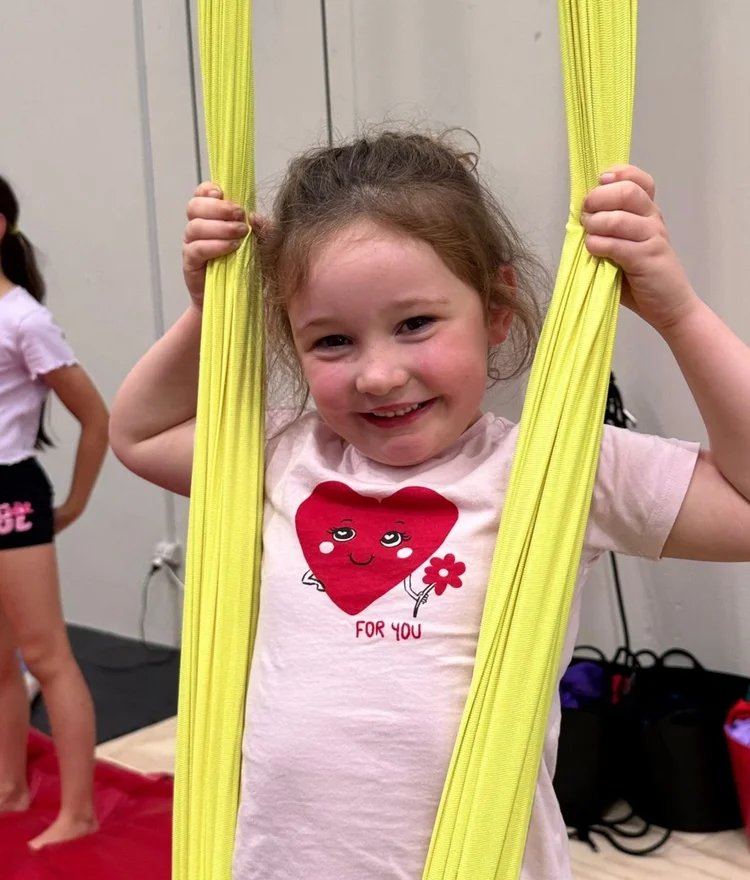 A grinning young girl holding yellow aerial silks in a kids class at the Grovedale studio