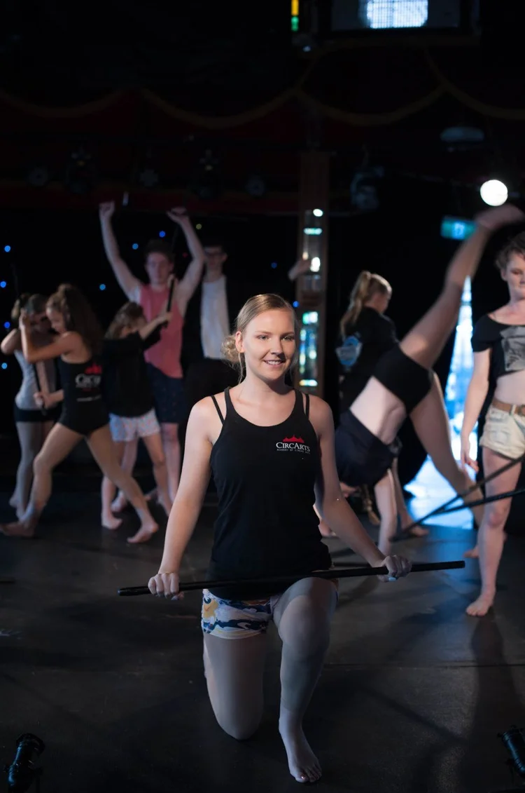A smiling dancer kneeling with a dance stick in a movement rehearsal while the group dances behind her