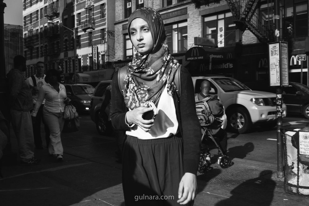 A young woman in a floral hijab walks into hard low sun on a Lower Manhattan pavement, looking straight into the lens; McDonald's signage and fire escapes behind, a stroller and long cast shadows at right