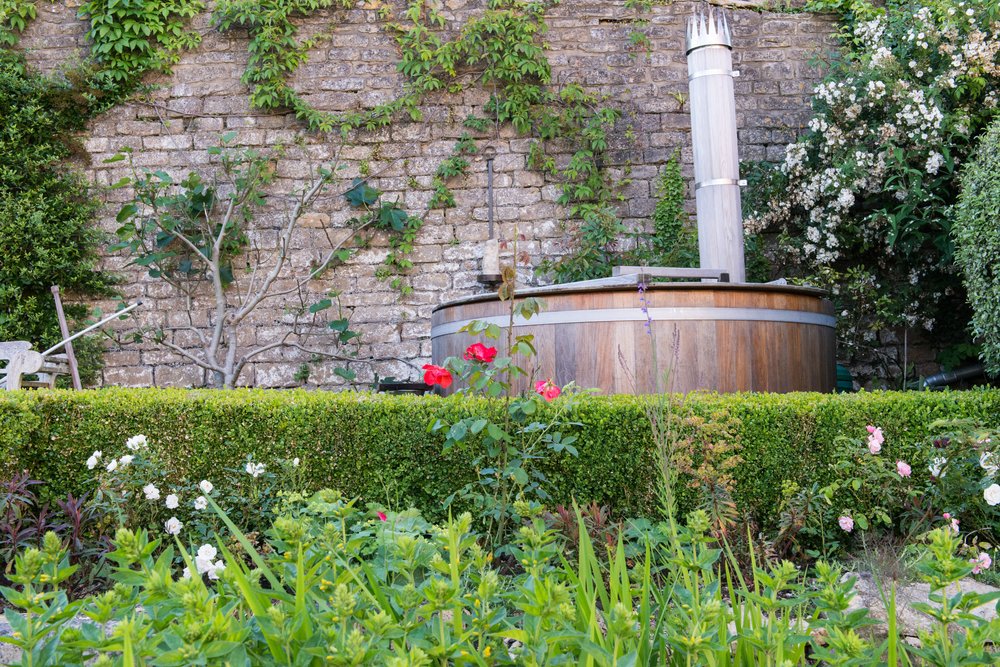 Wooden hot tub against an old stone wall behind a low clipped hedge and roses