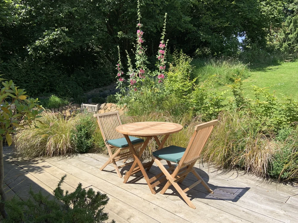 Bistro table on decking among grasses and pink hollyhocks