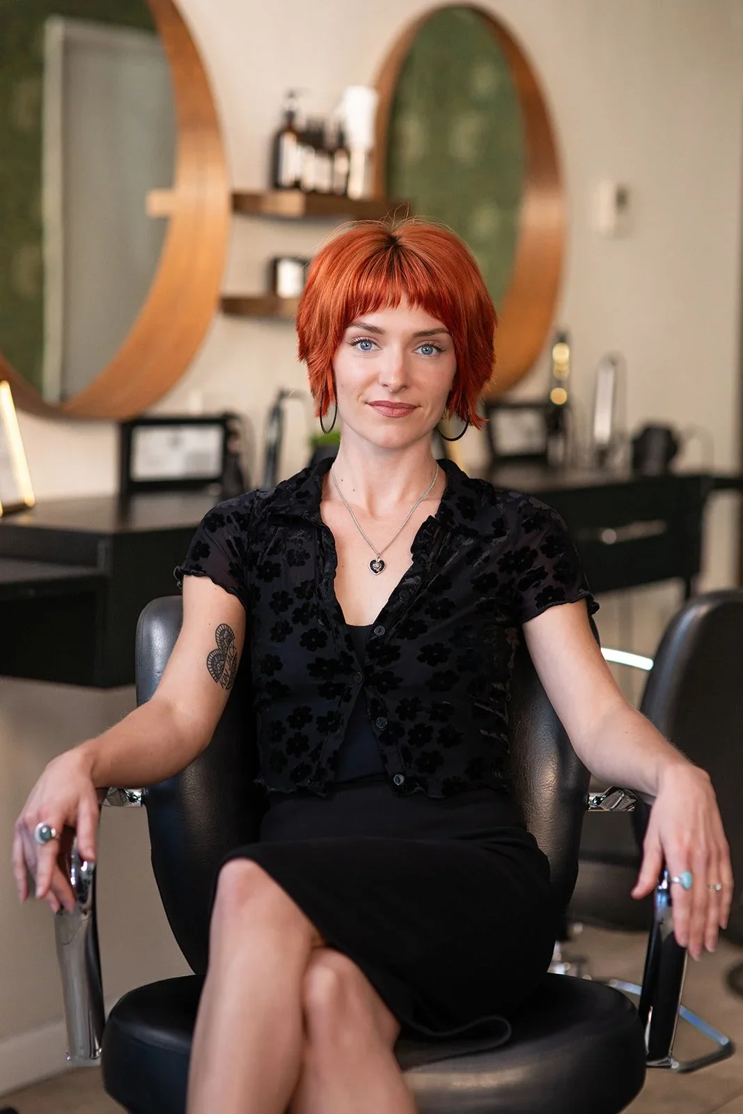 Taylor- A woman with short red hair and blue eyes sitting in a salon chair in, wearing a black floral velvety top and a skirt, with jewelry including rings, rings, and a pendant necklace.