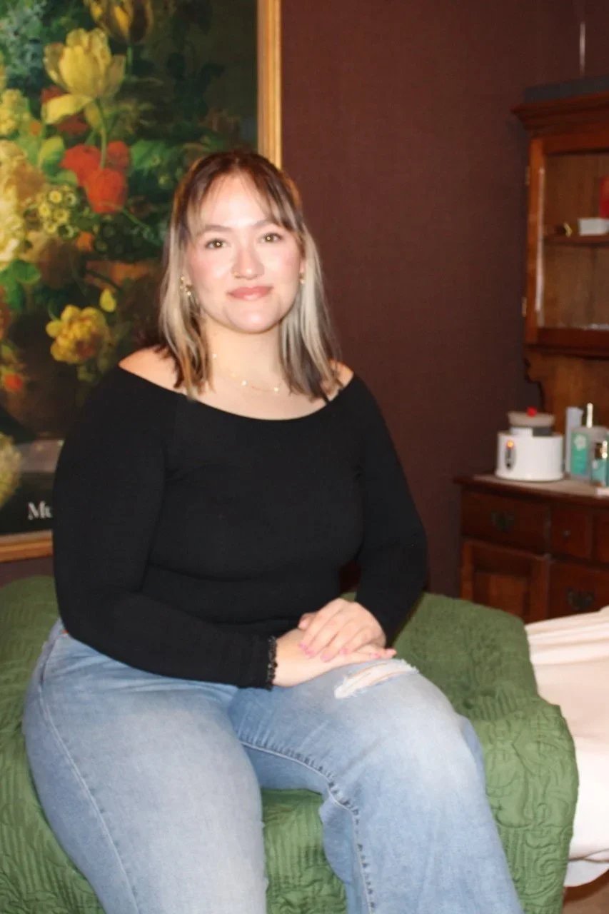 Juli- A woman with shoulder-length brown hair with blonde highlights, wearing a black long-sleeve top and light blue jeans, sitting in our treatment room.