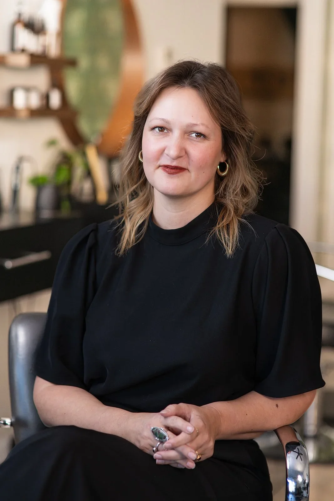 Melissa- A woman with wavy shoulder-length brown hair with blonde highlights wearing a black dress and gold hoop earrings, sitting the salon.