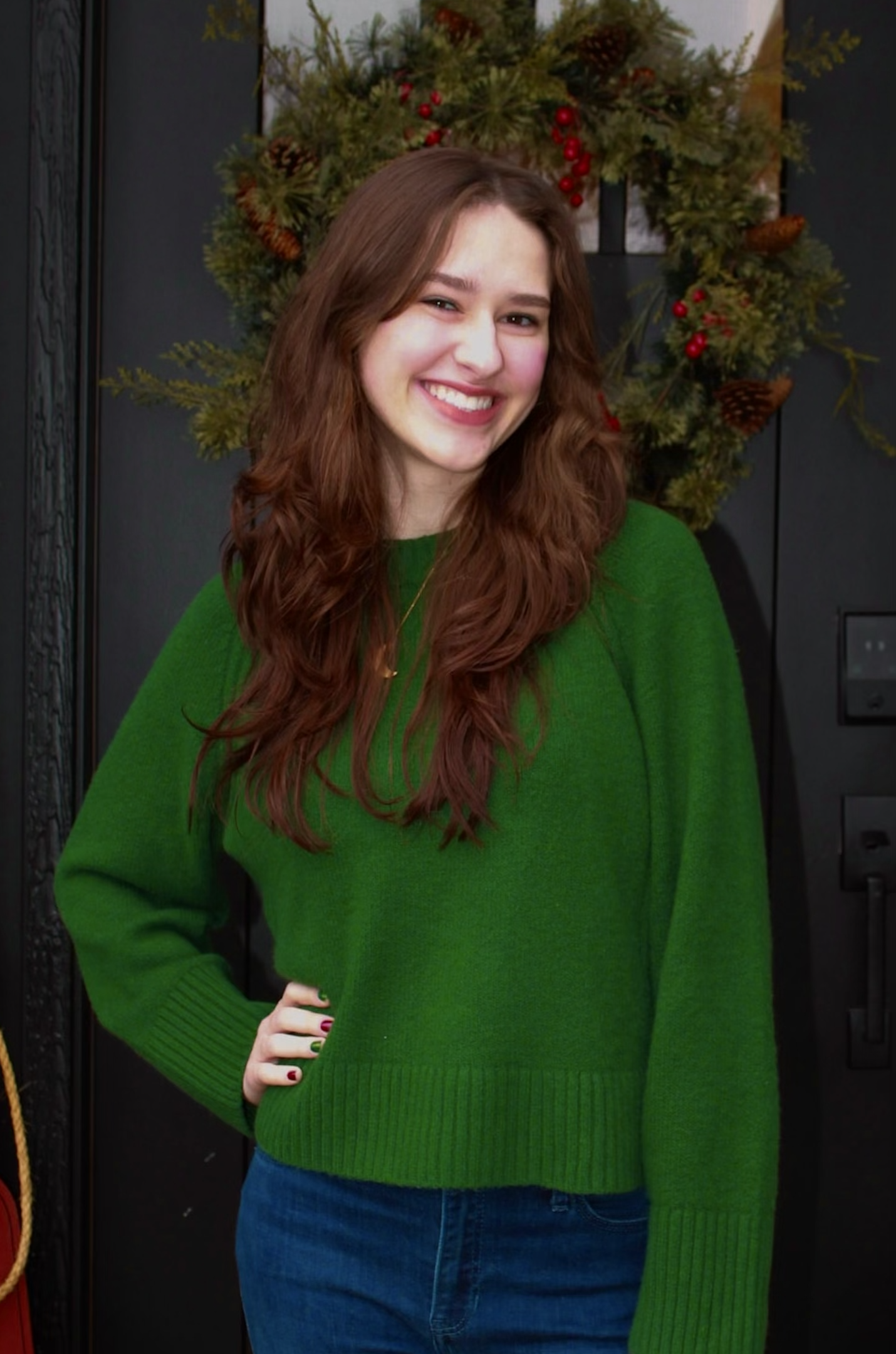 Natalie- A woman with long, wavy brown hair wearing a green sweater, standing in front of a door.