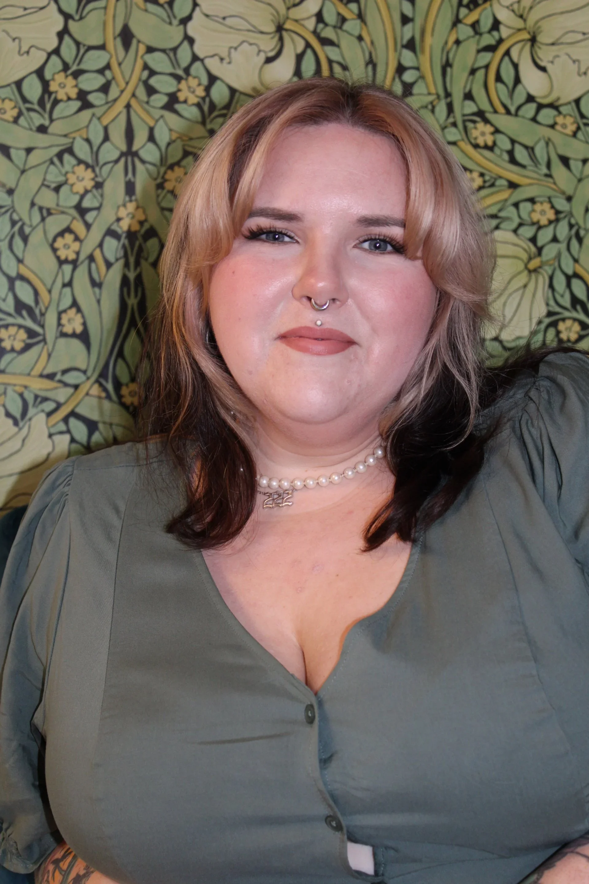 Christine- A woman with shoulder-length brown hair and a money piece, wearing a septum ring, a pearl necklace, and a green top, sitting in front of a decorative floral wallpaper background.