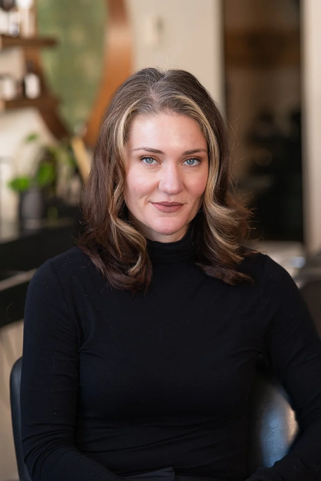 Easton- A woman with wavy shoulder-length brown hair, blue eyes, and fair skin, wearing a black turtleneck, sitting in the salon.