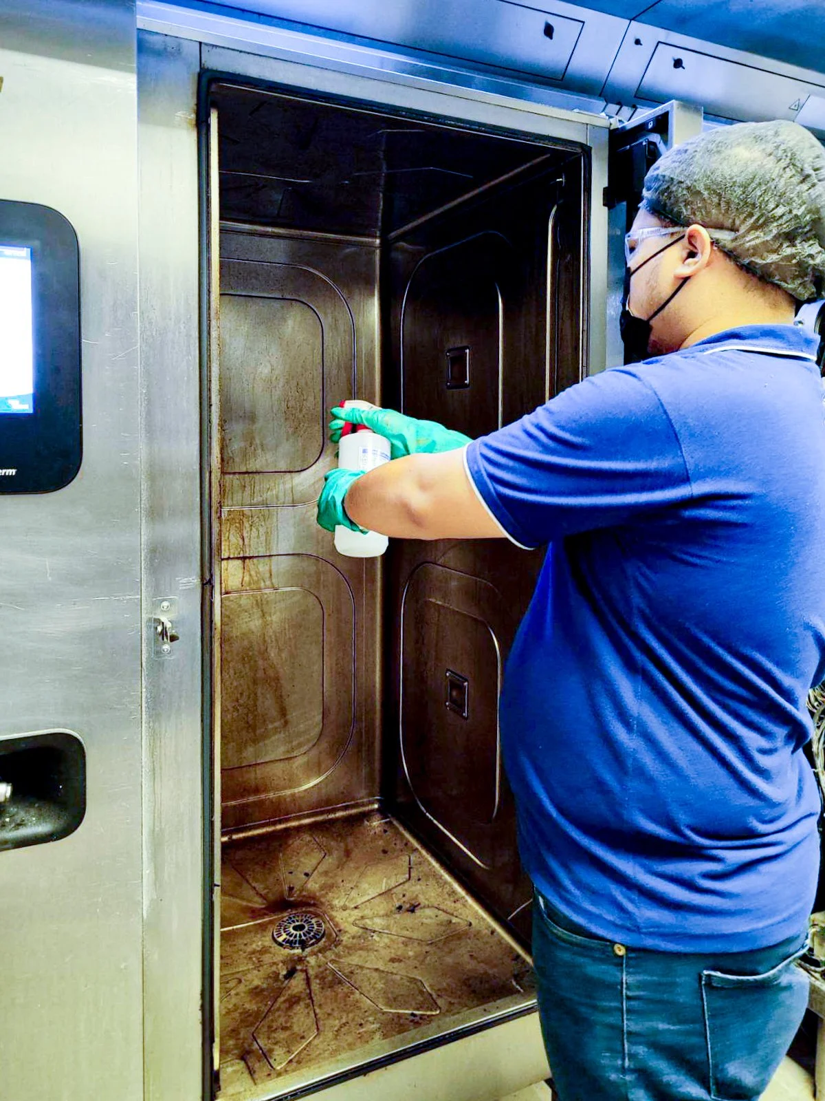 A person wearing a blue shirt, hair net, mask, and gloves cleaning the interior of an autoclave or sterilizer with a spray bottle.