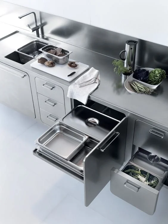 A modern stainless steel kitchen setup with a sink, a cutting board with shellfish, a towel, trays, and fresh vegetables on the countertop.