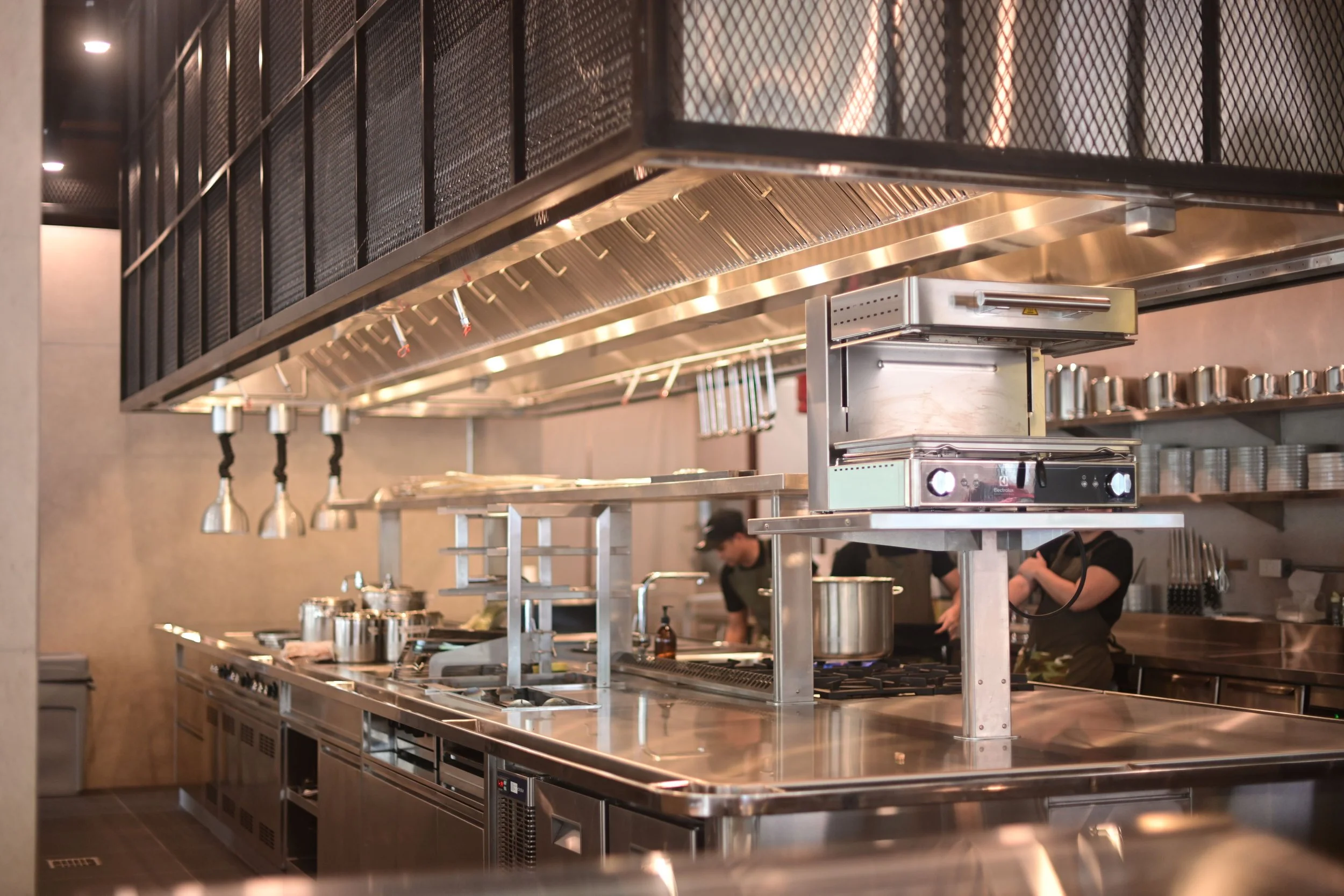 Modern stainless steel commercial kitchen with chefs preparing food, hanging lights, and metal shelving.