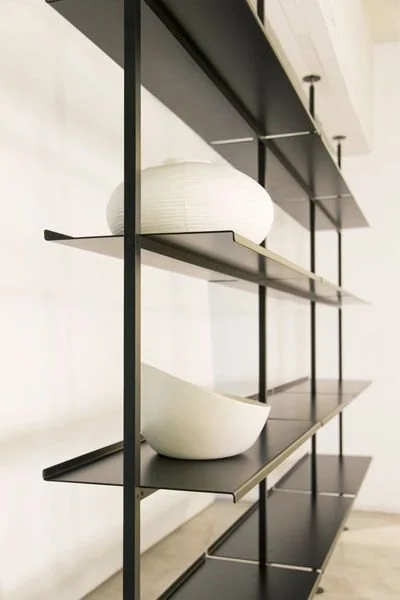 Black metal shelving unit with three shelves, holding a round white vase and a white bowl, against a white wall.