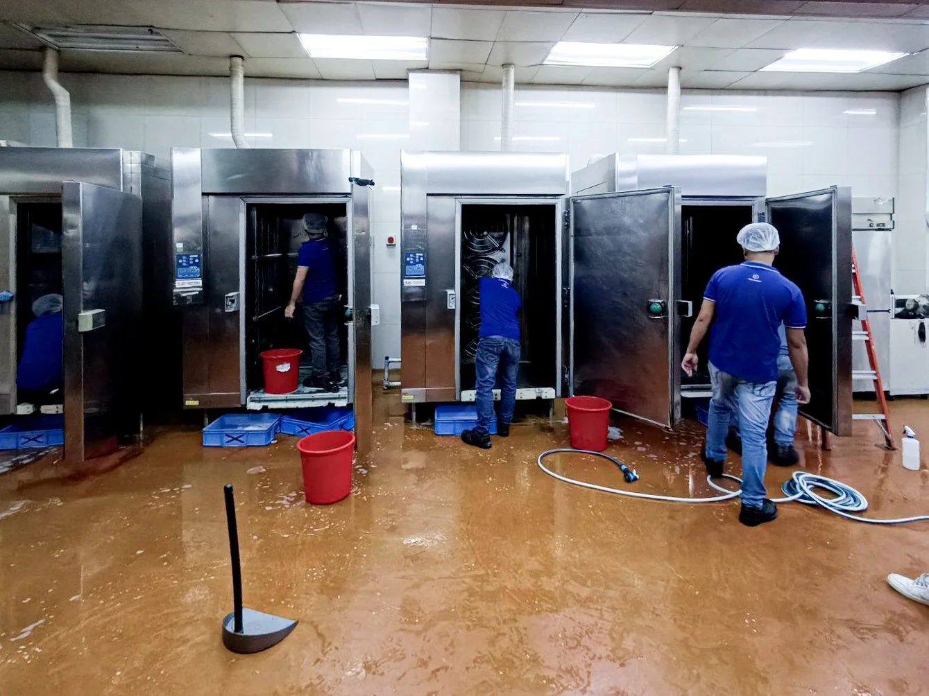 Workers cleaning industrial dishwashers in a commercial kitchen, with cleaning supplies and equipment around.