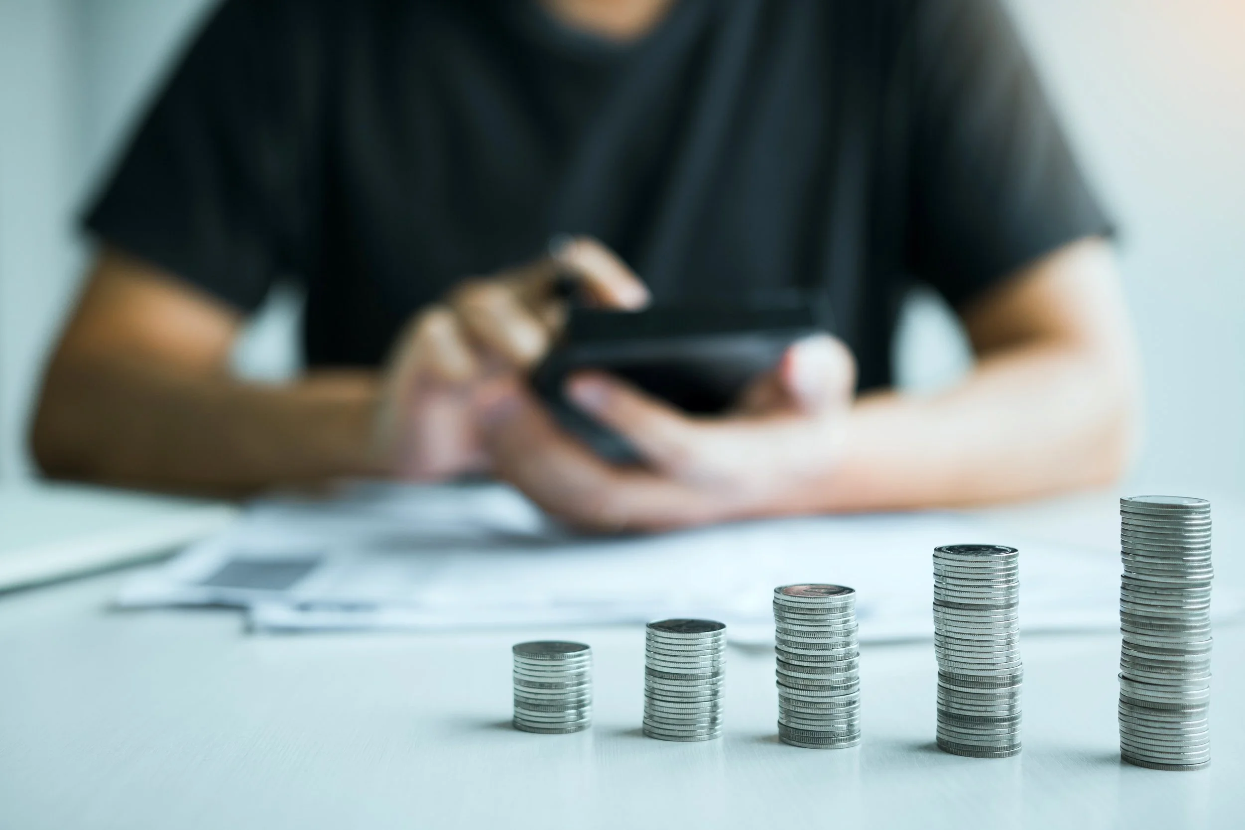 Person in black shirt sitting at desk, using a smartphone. In front of them, there are stacks of coins arranged in increasing height from left to right.