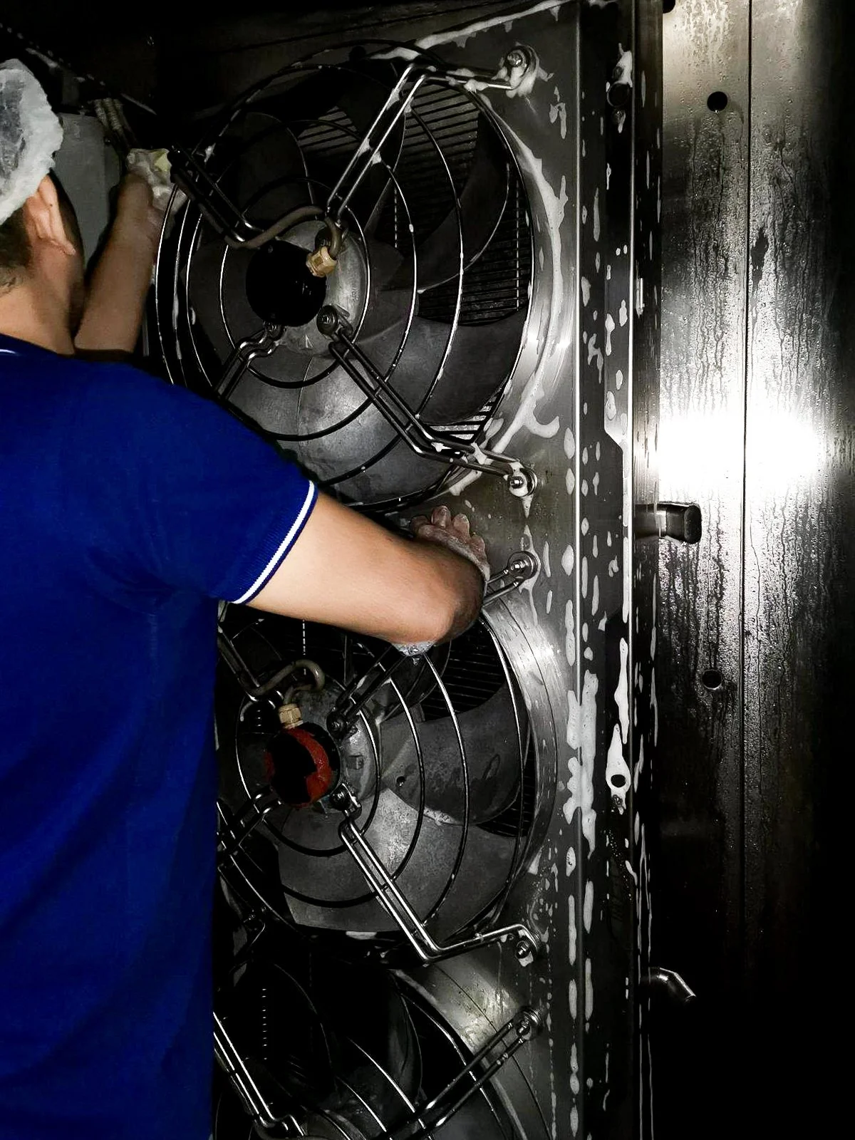 A person cleaning or repairing industrial stainless steel equipment with three large, round, winged fans or turbines, in a sanitary or food processing environment.