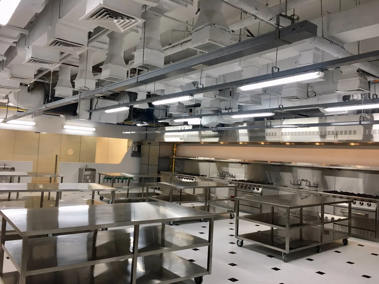 Empty commercial kitchen with stainless steel counters, gas stoves, and ventilation ducts on the ceiling.