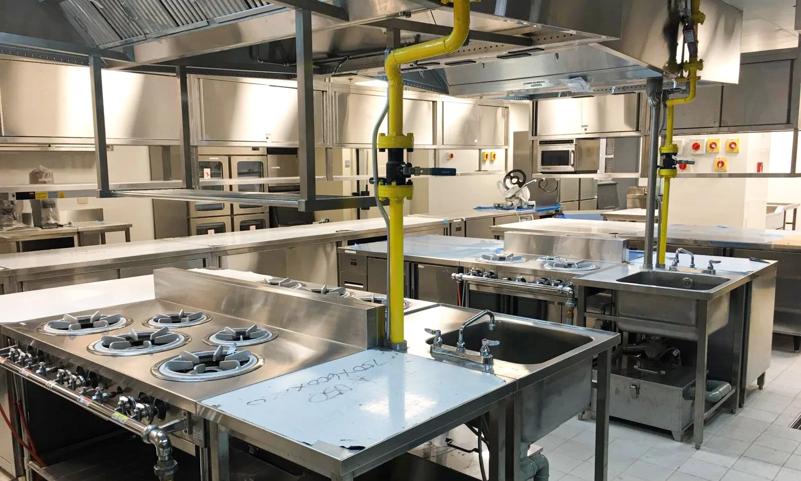Empty commercial kitchen with stainless steel appliances, sinks, ovens, and gas burners, with yellow pipes and electrical panels on walls.