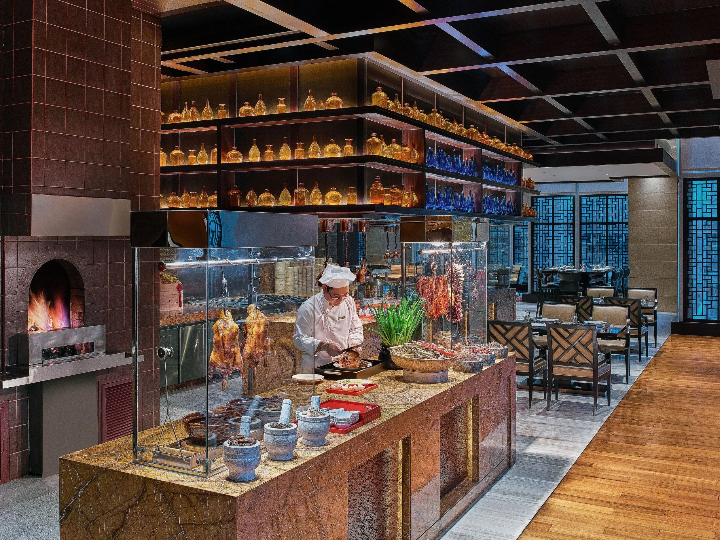 Restaurant kitchen with a chef preparing food at a marble counter, hanging roasted chickens, shelves with glass bottles and decorations, and dining area in the background.