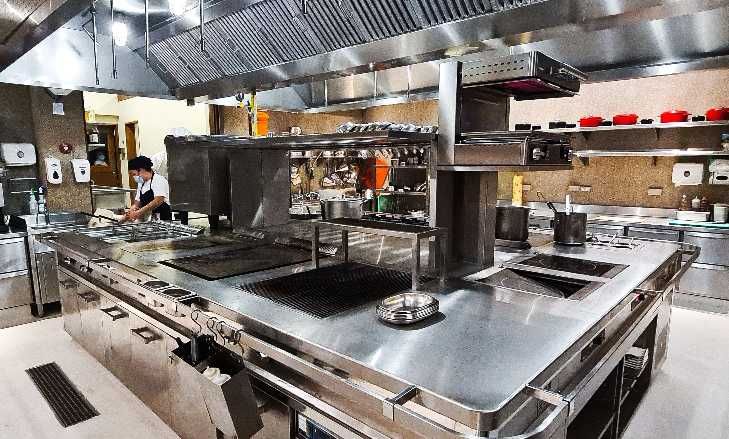 Commercial kitchen with stainless steel countertops, stove, pots, and a person wearing a mask and apron preparing food.