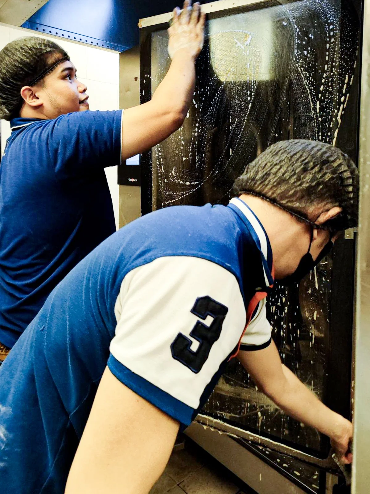 Two people cleaning a large glass oven door with soap suds and scrub brushes.