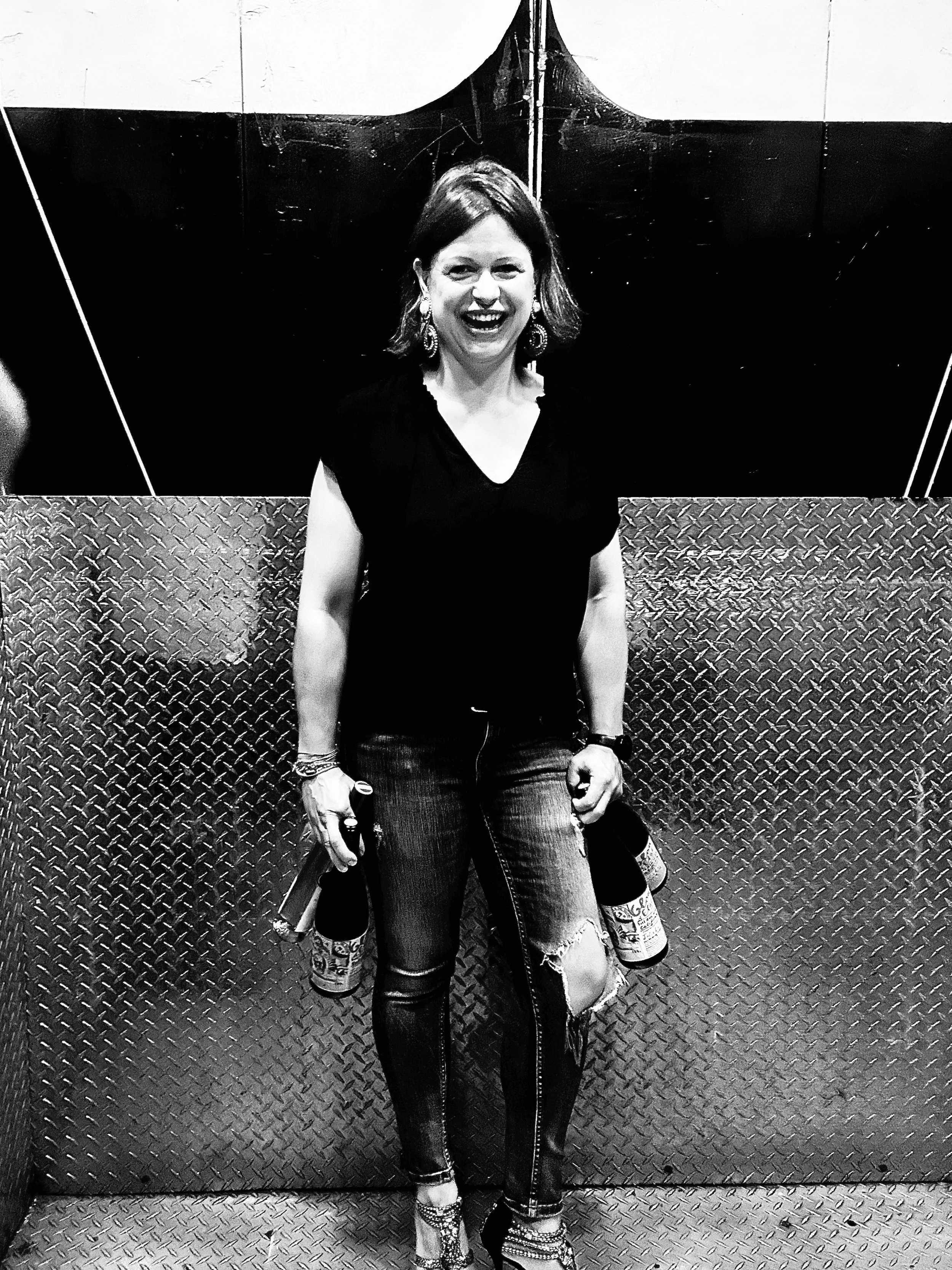 Katja is wearing a black v neck t-shirt, holding two wine bottles in each hand