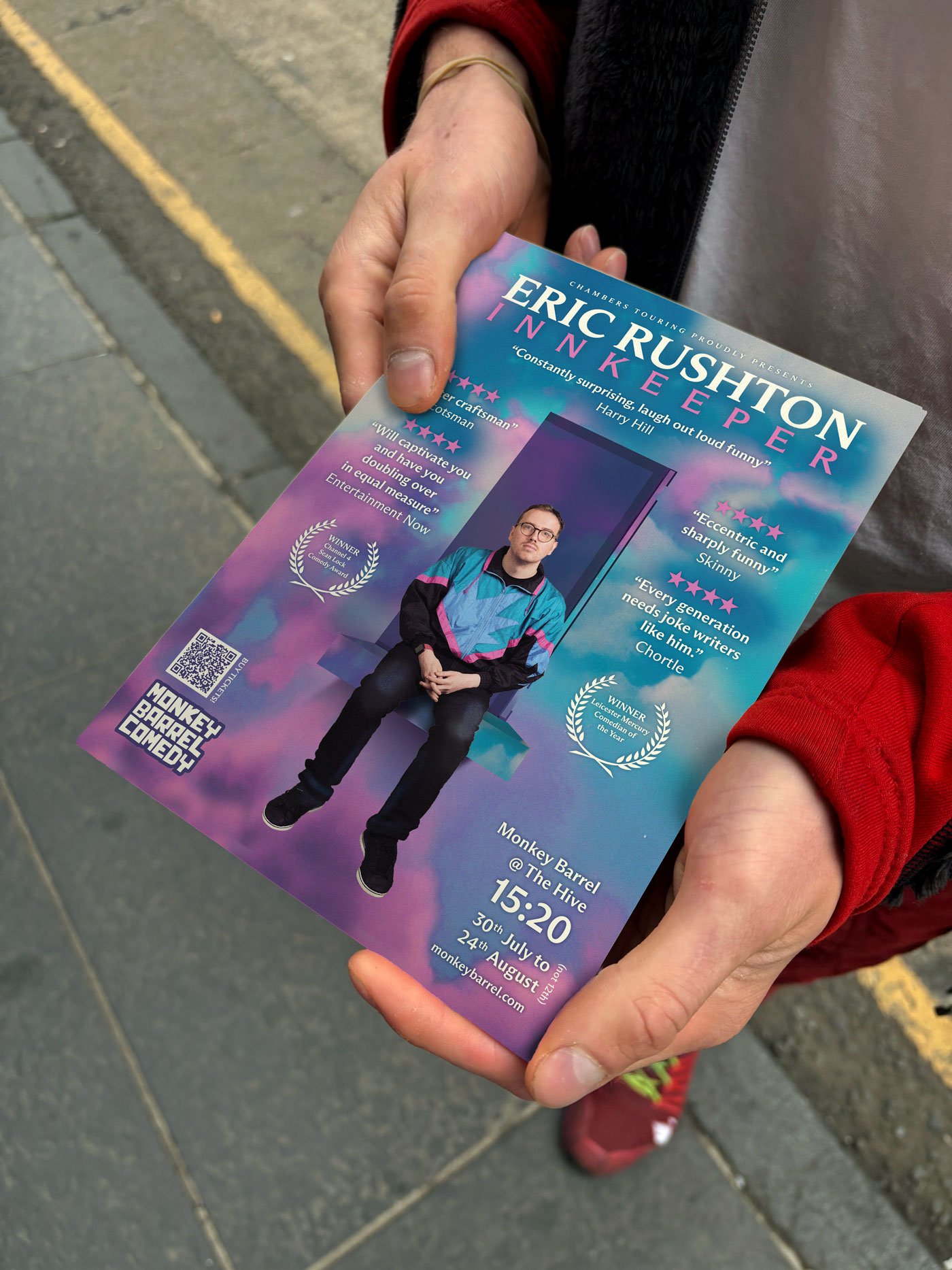 A person holding a colorful flyer for Erich Rushton, an entertainer, featuring a photo of him sitting on a bench wearing glasses and a retro jacket. The flyer includes event details at Monkey Barrel, The Hive, from July 30th to August 24th at 15:20.