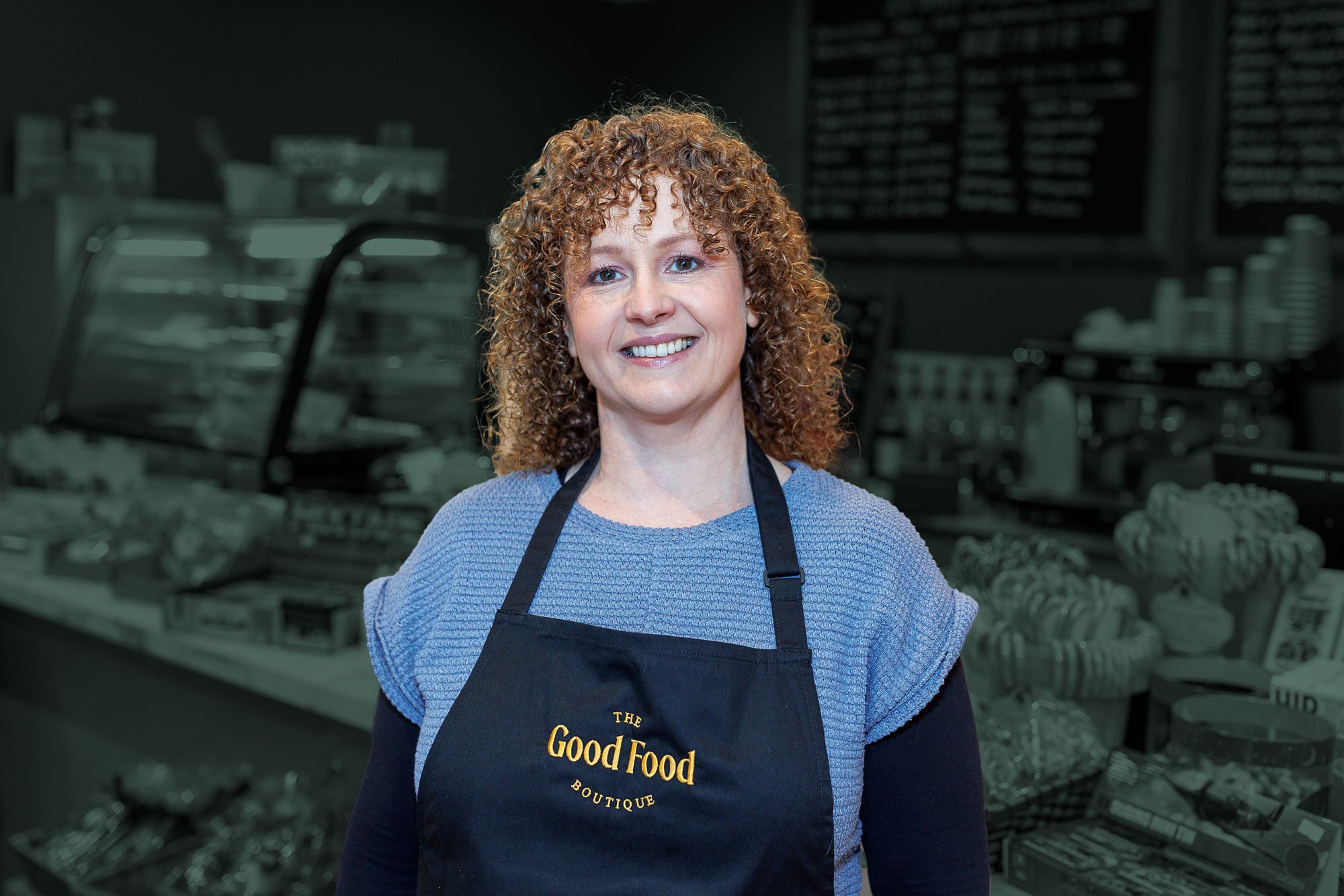 A woman with curly red hair smiling while wearing a blue shirt and a black apron with the text 'The Good Food Boutique' in a bakery or food shop.