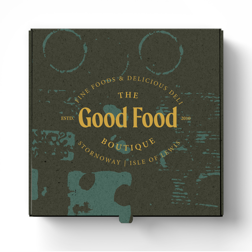 A dark-colored box labeled 'The Good Food' from 'The Good Food Boutique' with gold lettering, featuring a vintage design and text indicating it is from Tornoway, Isle of Lewis, established in 2010.