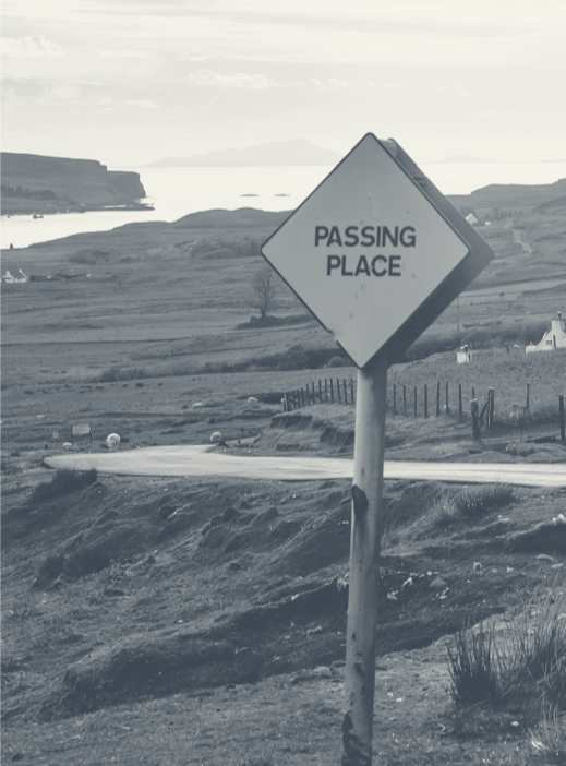 A rural road with a diamond-shaped sign that reads 'Passing Place' in black letters, with rolling hills and body of water in the background.
