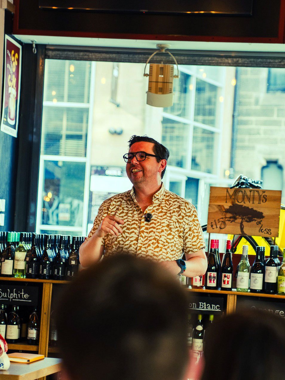 A man with glasses and a patterned shirt speaking to an audience inside a wine shop or tasting room, with bottles of wine on display behind him and a window showing a city street outside.