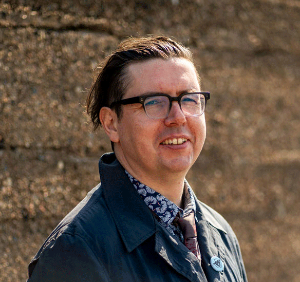 A man with glasses and slicked-back hair standing outdoors in front of a rocky background.