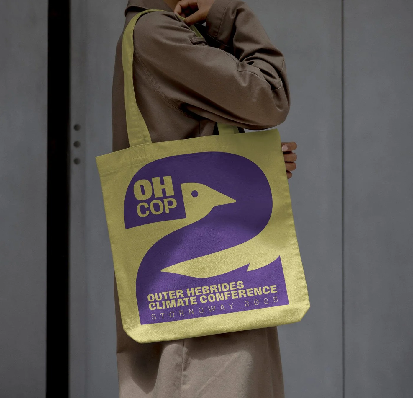Person holding a yellow tote bag with purple text and a duck logo at the Outer Hebrides Climate Conference, Stormaway 2025, standing in front of a metallic wall.