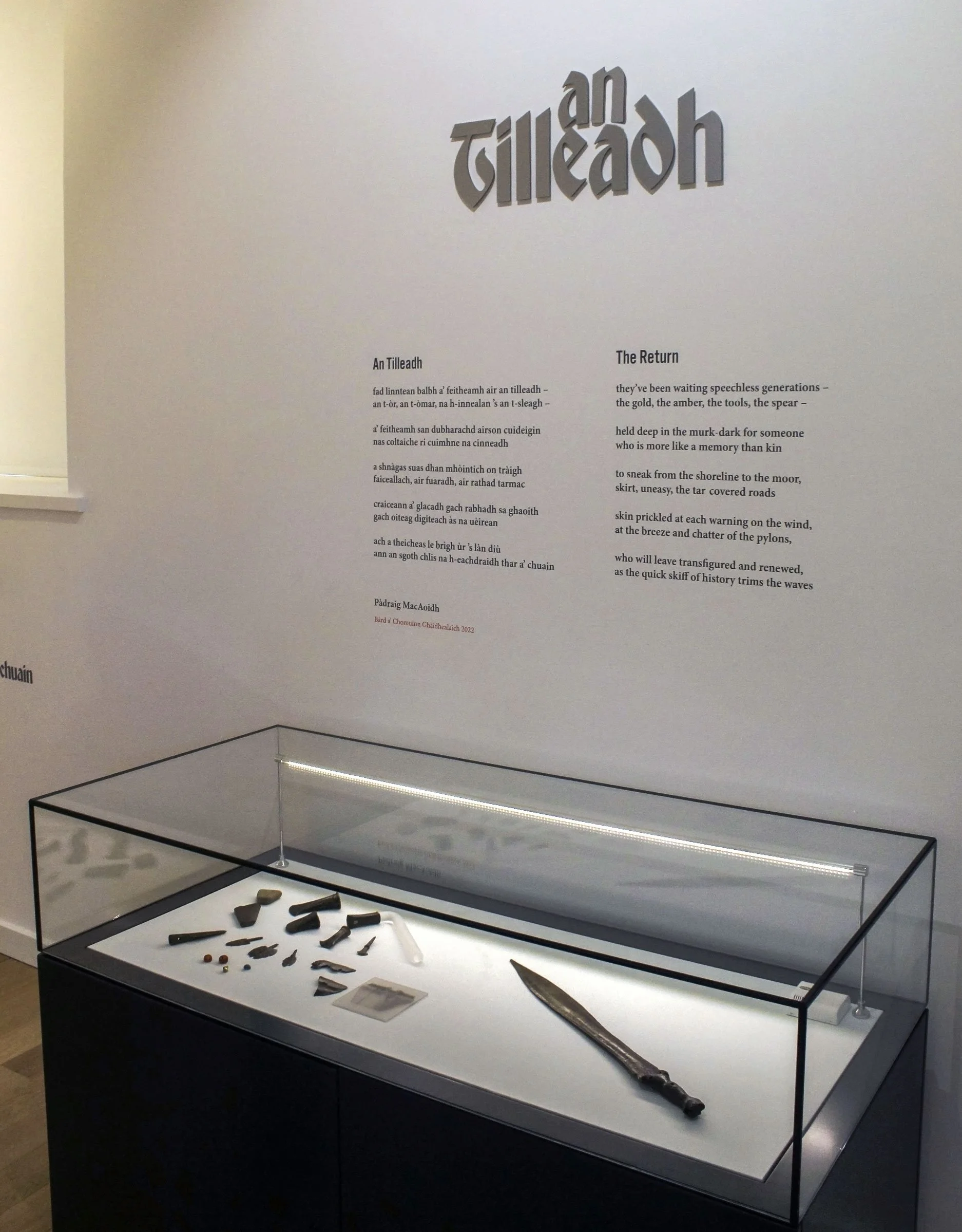 Museum display case with ancient artifacts and a large knife, with descriptive text on the wall in a different language and English.