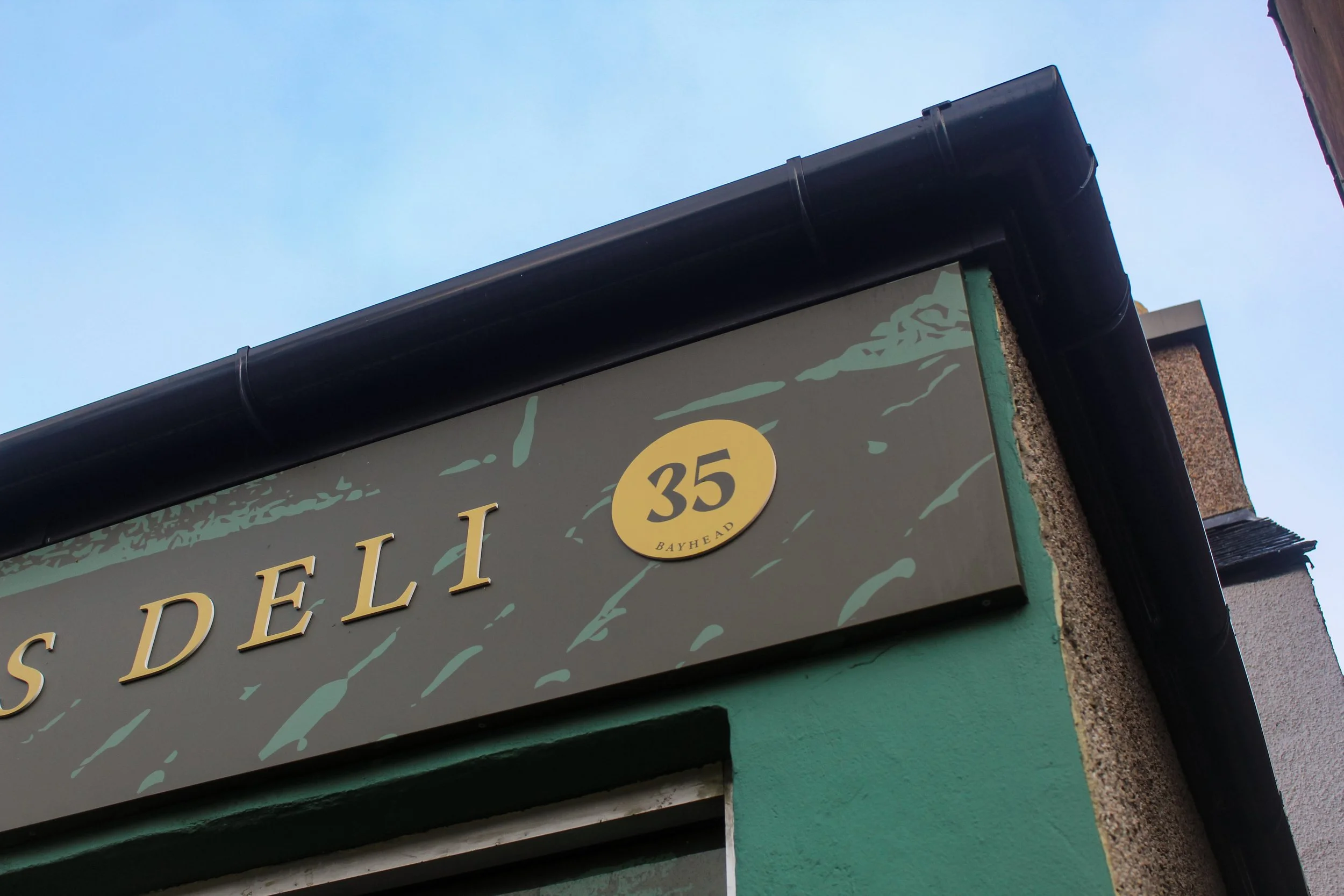 Street corner sign showing '35 Bayhead' above the partially visible word 'S Deli' on a green building with a black gutter and blue sky in the background.