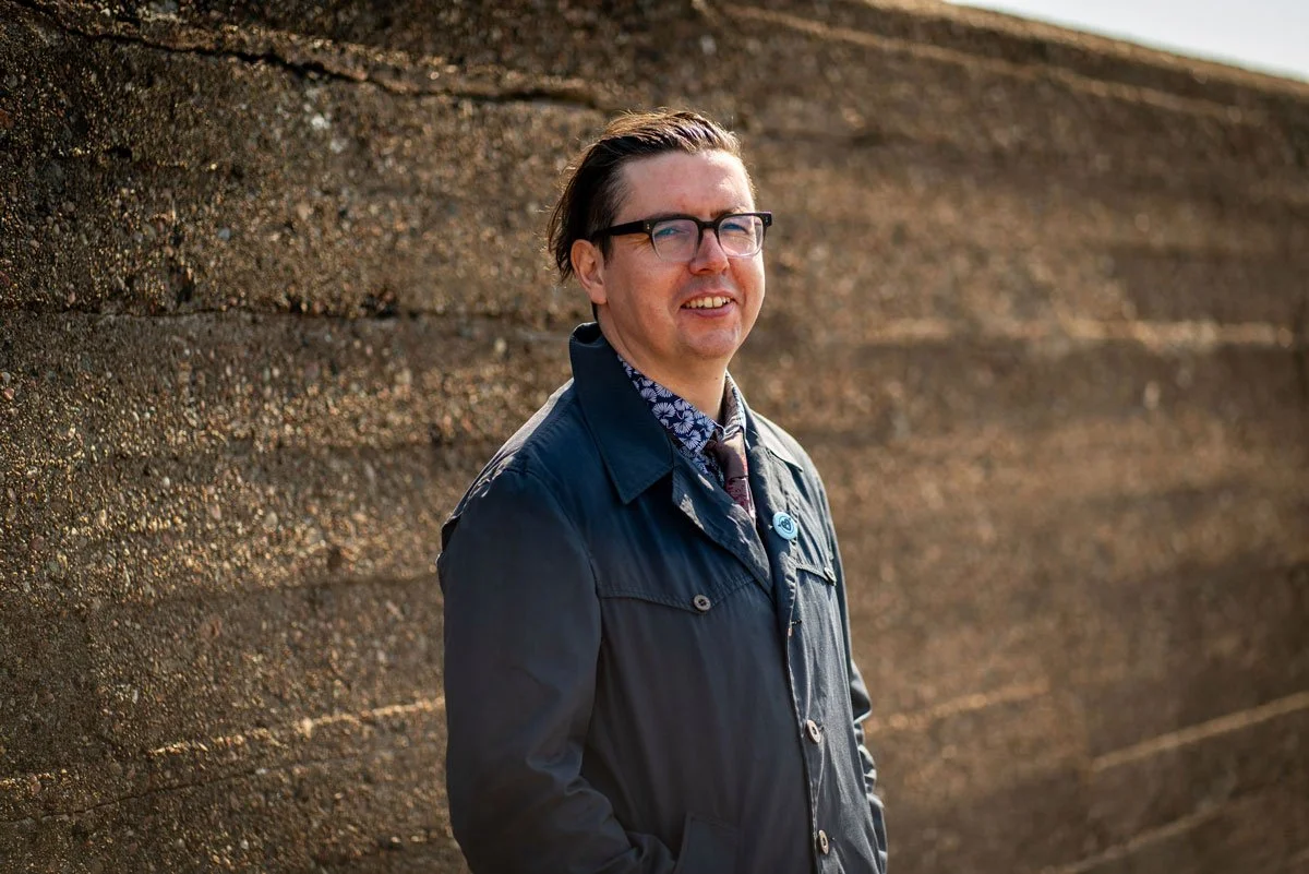 A man with glasses standing outdoors next to a textured concrete wall, wearing a dark jacket and patterned shirt.